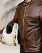 Person wearing a brown leather jacket holding a white helmet with black and orange stripes.